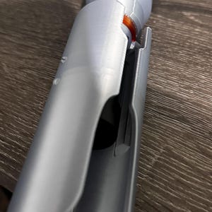 Puede incluir: Primer plano de una empu&ntilde;adura de sable de luz plateada con un dise&ntilde;o detallado. La empu&ntilde;adura est&aacute; hecha de un material impreso en 3D y presenta una secci&oacute;n que se desliza, revelando un interior naranja. El sable de luz est&aacute; sobre una superficie de vetas de madera oscura.