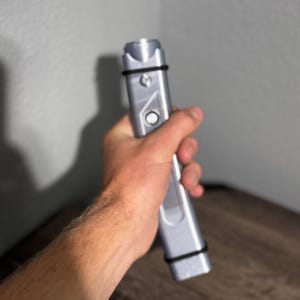 Pu&ograve; includere: Un dispositivo rettangolare color argento con una parte superiore cilindrica e un elastico nero avvolto attorno. Il dispositivo ha un pulsante e un design geometrico. L'oggetto &egrave; tenuto in mano da una persona.