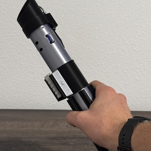 Pu&ograve; includere: Un'elica di spada laser nera, argento e grigia. L'elica ha un corpo cilindrico con un'impugnatura nera e una sezione centrale argentata. La parte superiore dell'elica ha un design nero angolato. L'elica &egrave; tenuta in una mano.