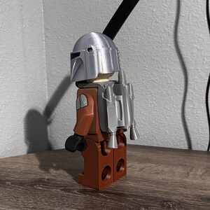 May include: A plastic toy figure featuring a silver helmet, a brown torso and legs, black hands, and a silver jetpack attached to the back. A long, thin, black rectangular piece extends upward from the back of the helmet, resembling a blade.