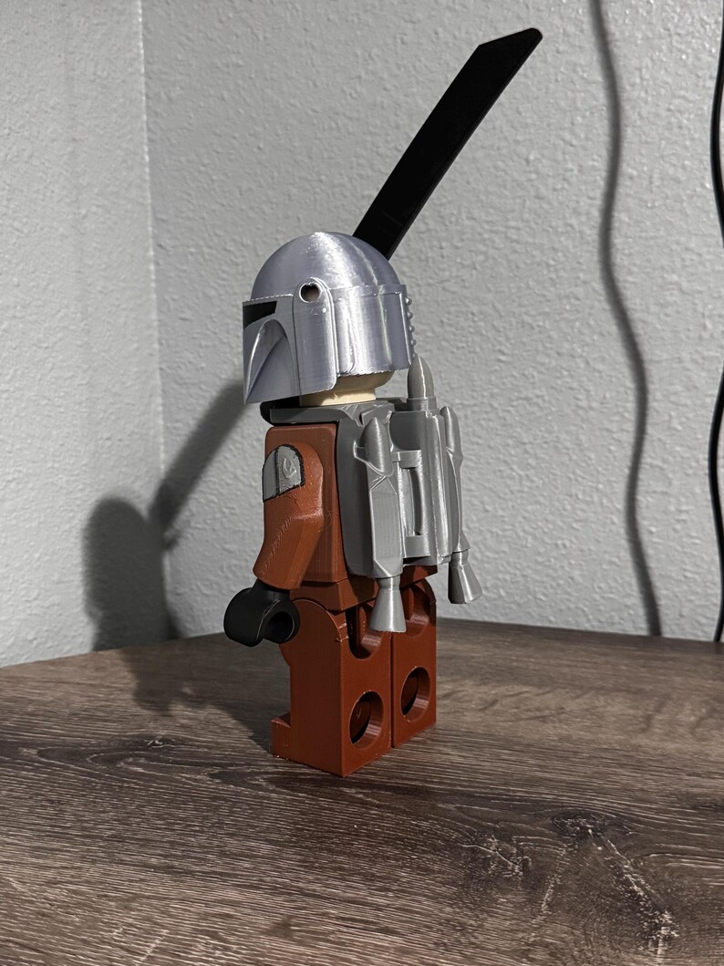 May include: A plastic toy figure featuring a silver helmet, a brown torso and legs, black hands, and a silver jetpack attached to the back. A long, thin, black rectangular piece extends upward from the back of the helmet, resembling a blade.
