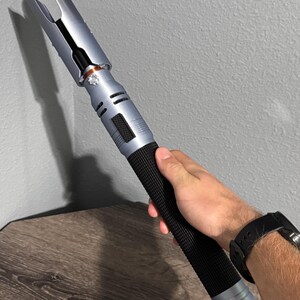 Puede incluir: Un accesorio de sable de luz plateado y negro con un mango negro texturizado. El sable de luz tiene un dise&ntilde;o &uacute;nico con un emisor bifurcado y lo sostiene una persona. El sable de luz mide aproximadamente 30 cm de largo.