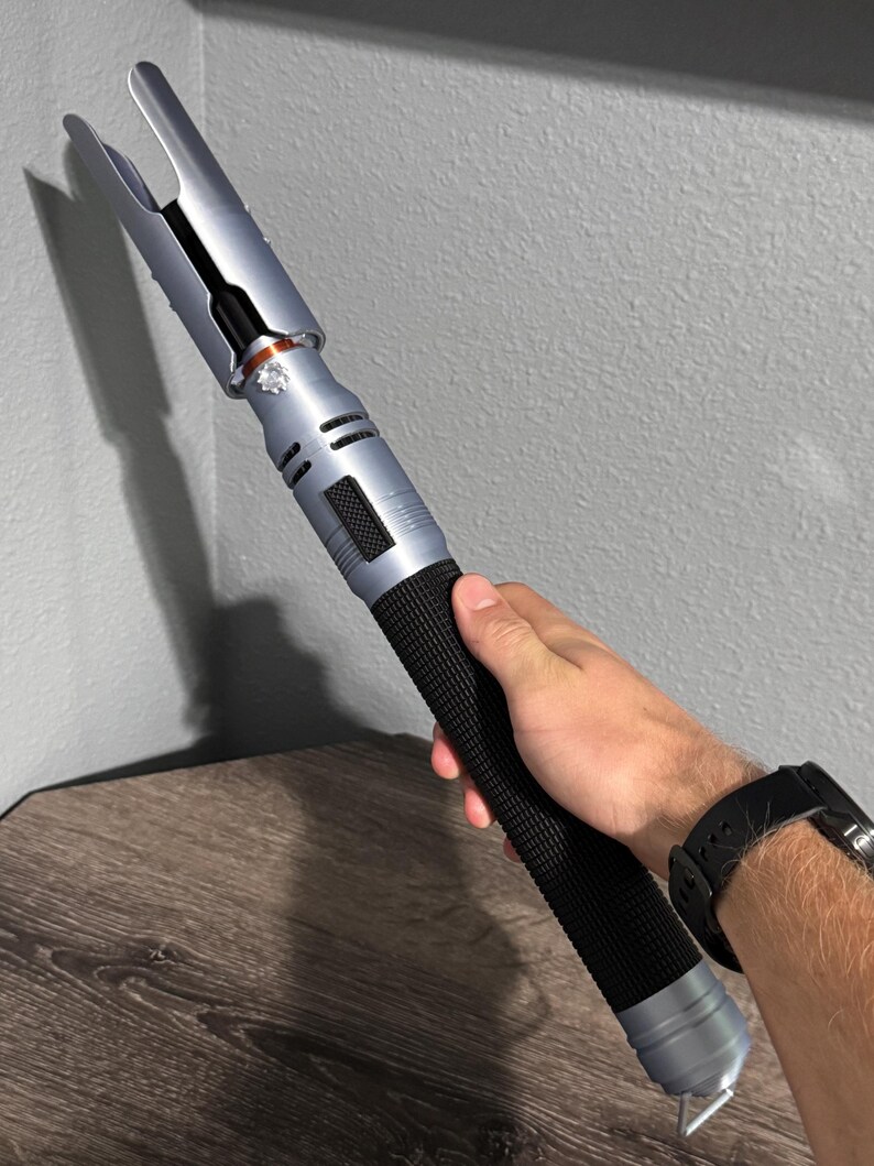 Puede incluir: Un accesorio de sable de luz plateado y negro con un mango negro texturizado. El sable de luz tiene un dise&ntilde;o &uacute;nico con un emisor bifurcado y lo sostiene una persona. El sable de luz mide aproximadamente 30 cm de largo.