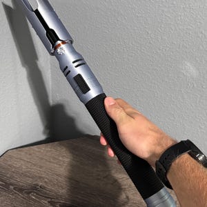 Puede incluir: Un sable de luz plateado y negro con un mango negro texturizado. La parte superior del sable de luz tiene un dise&ntilde;o &uacute;nico con dos p&uacute;as. El sable de luz se sostiene contra una pared gris y una superficie de madera.