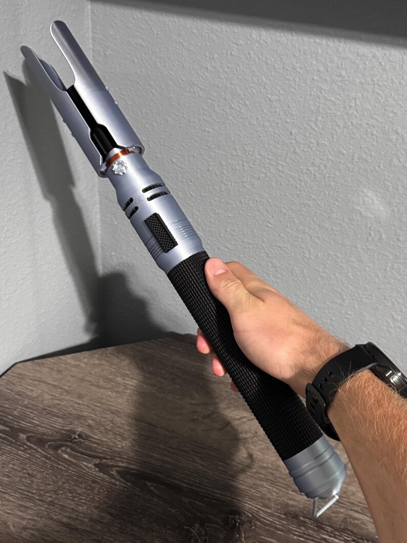 Puede incluir: Un sable de luz plateado y negro con un mango negro texturizado. La parte superior del sable de luz tiene un dise&ntilde;o &uacute;nico con dos p&uacute;as. El sable de luz se sostiene contra una pared gris y una superficie de madera.