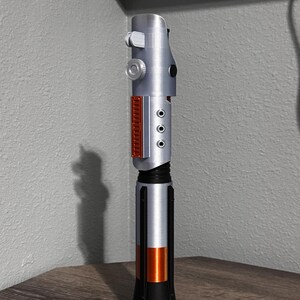 May include: A silver and black lightsaber prop with orange and red accents. The hilt has a detailed design with buttons and panels. The lightsaber is standing upright on a wooden surface.