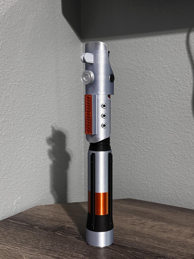 May include: A silver and black lightsaber prop with orange and red accents. The hilt has a detailed design with buttons and panels. The lightsaber is standing upright on a wooden surface.