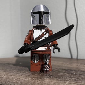 May include: A brown and white plastic toy figure wearing a silver helmet and holding a black sword. A smaller, matching miniature figure stands at the feet of the larger figure. Both figures feature detailed armor patterns and black hand pieces.