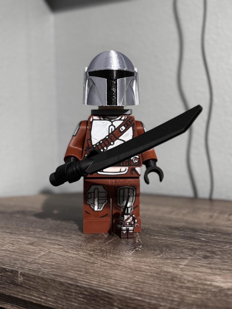May include: A brown and white plastic toy figure wearing a silver helmet and holding a black sword. A smaller, matching miniature figure stands at the feet of the larger figure. Both figures feature detailed armor patterns and black hand pieces.