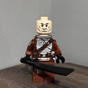 May include: A plastic minifigure featuring a light skin tone head with a printed beard and mustache. The torso and legs are brown with silver armor plating details. The figure has black hands and is holding a long, black, flat-bladed sword.