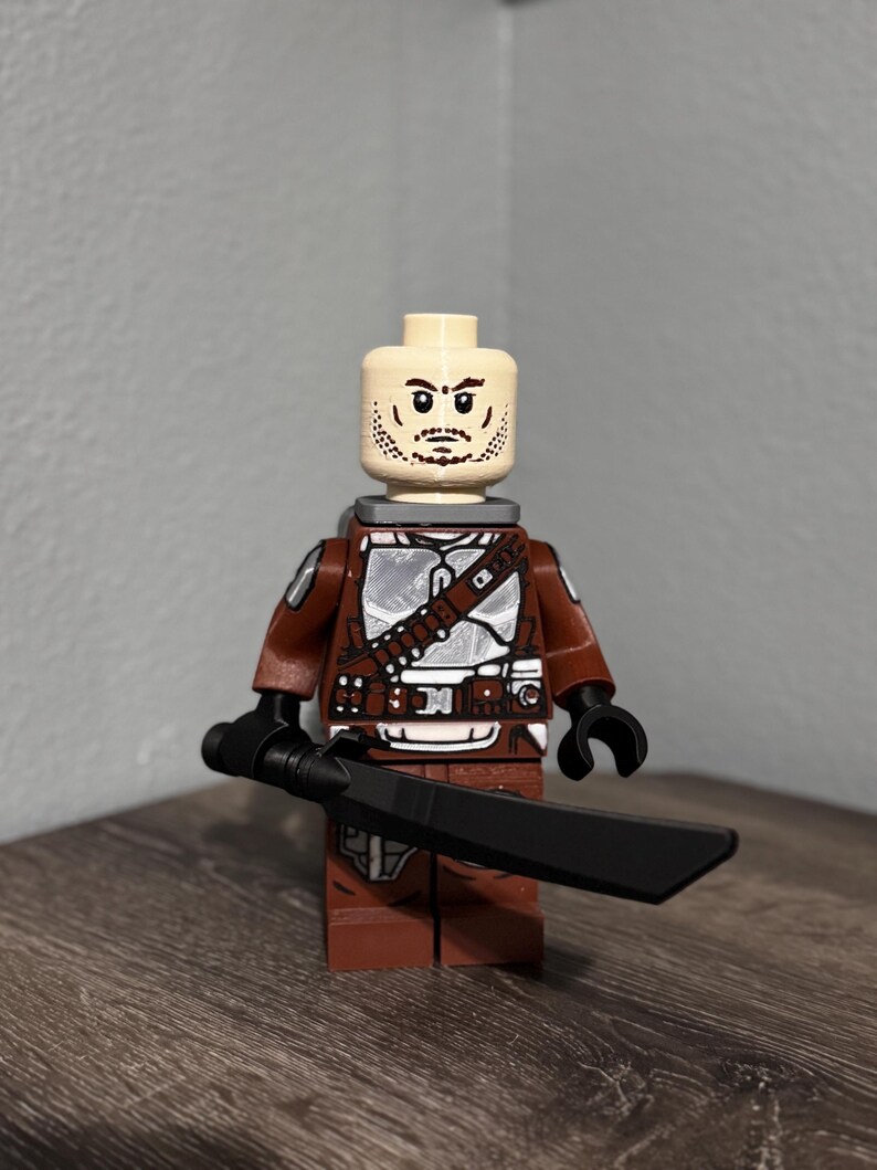 May include: A plastic minifigure featuring a light skin tone head with a printed beard and mustache. The torso and legs are brown with silver armor plating details. The figure has black hands and is holding a long, black, flat-bladed sword.