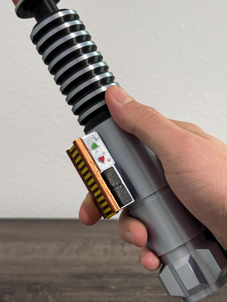 Puede incluir: Un accesorio de sable de luz plateado y negro con un mango detallado y un panel de control con botones rojos y verdes. El accesorio se sostiene en una mano, mostrando su dise&ntilde;o y construcci&oacute;n.