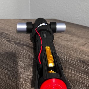 May include: Black and silver lightsaber with a red emitter and gold accents. The hilt is black with silver details and a red wire. The emitter is a bright red circle. The lightsaber is on a wooden surface.