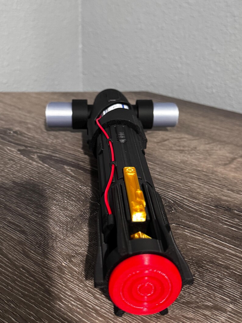 May include: Black and silver lightsaber with a red emitter and gold accents. The hilt is black with silver details and a red wire. The emitter is a bright red circle. The lightsaber is on a wooden surface.