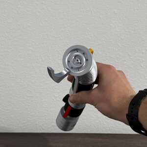 May include: A silver and black lightsaber prop with a red accent, held in a person's hand. The lightsaber features a detailed hilt and a circular emitter. The background is a plain white wall and a dark wooden surface.
