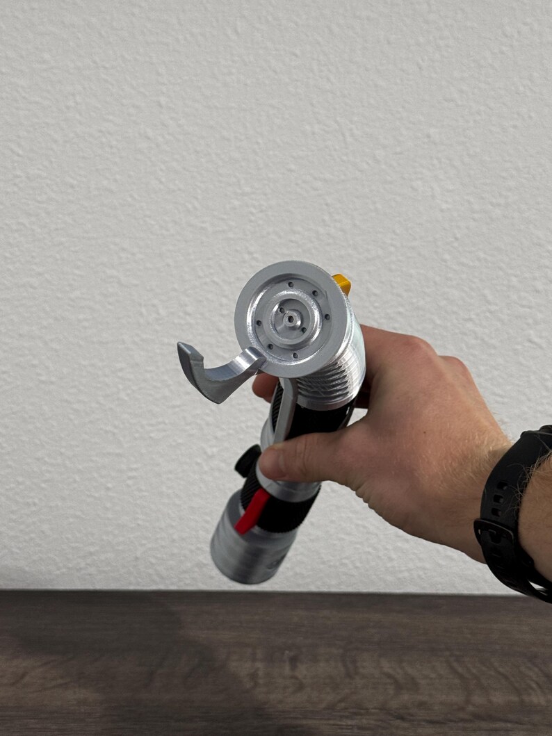 May include: A silver and black lightsaber prop with a red accent, held in a person's hand. The lightsaber features a detailed hilt and a circular emitter. The background is a plain white wall and a dark wooden surface.
