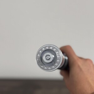 Puede incluir: Primer plano de un molinillo de pimienta plateado y negro. La parte superior tiene un dise&ntilde;o circular con peque&ntilde;os agujeros. El molinillo se sostiene en una mano sobre un fondo neutro.