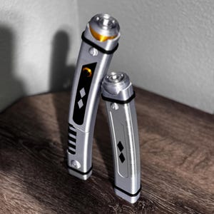 Pu&ograve; includere: Due spade laser argentate con dettagli neri e dorati. Le spade laser hanno un design elegante e metallico con fasce nere e intarsi a forma di diamante. Una &egrave; leggermente pi&ugrave; alta dell'altra, entrambe sono appoggiate su una superficie di legno.