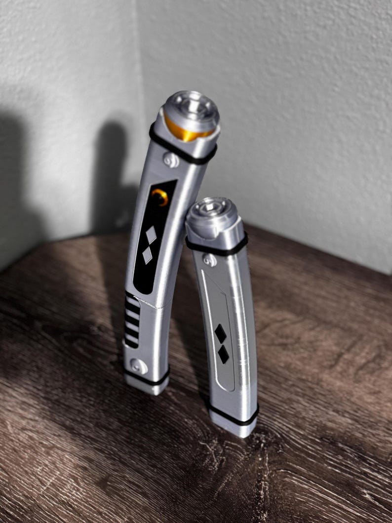 Pu&ograve; includere: Due spade laser argentate con dettagli neri e dorati. Le spade laser hanno un design elegante e metallico con fasce nere e intarsi a forma di diamante. Una &egrave; leggermente pi&ugrave; alta dell'altra, entrambe sono appoggiate su una superficie di legno.