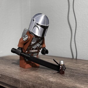 May include: A large brown and silver plastic Mandalorian minifigure wearing a metallic silver helmet and holding a long black sword. The sword tip rests on a much smaller, miniature version of the same character, also featuring a silver helmet.