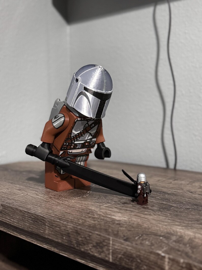 May include: A large brown and silver plastic Mandalorian minifigure wearing a metallic silver helmet and holding a long black sword. The sword tip rests on a much smaller, miniature version of the same character, also featuring a silver helmet.