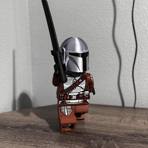 May include: A plastic toy figure resembling a space warrior wearing a metallic silver helmet and brown armor with silver accents. The figure holds a long, thin black sword-like weapon upright in one hand, standing in a forward-facing pose.