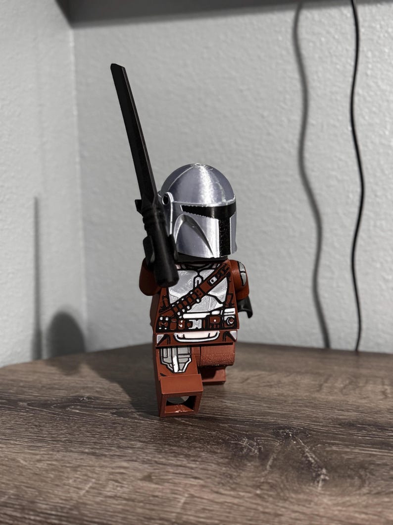 May include: A plastic toy figure resembling a space warrior wearing a metallic silver helmet and brown armor with silver accents. The figure holds a long, thin black sword-like weapon upright in one hand, standing in a forward-facing pose.