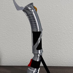 May include: A silver lightsaber with a curved blade and a black grip. The hilt features a red button and a yellow accent. The lightsaber is displayed on a black stand.