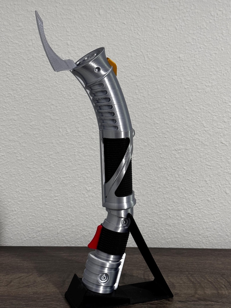 May include: A silver lightsaber with a curved blade and a black grip. The hilt features a red button and a yellow accent. The lightsaber is displayed on a black stand.