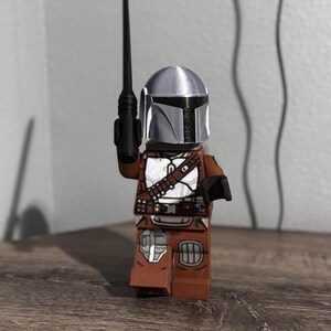 May include: A plastic toy figure resembling a space warrior wearing a metallic silver helmet with a black visor. The figure has a brown torso and legs with white armor plating details and a black belt. It holds a long, thin black weapon in one hand.