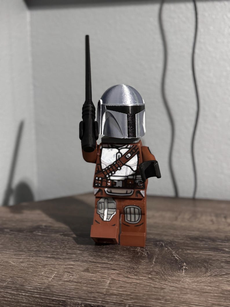 May include: A plastic toy figure resembling a space warrior wearing a metallic silver helmet with a black visor. The figure has a brown torso and legs with white armor plating details and a black belt. It holds a long, thin black weapon in one hand.