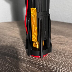 May include: A black and gold lightsaber hilt with red detailing. The hilt has a gold rectangular feature and a red wire. The base is encircled by a red ring.