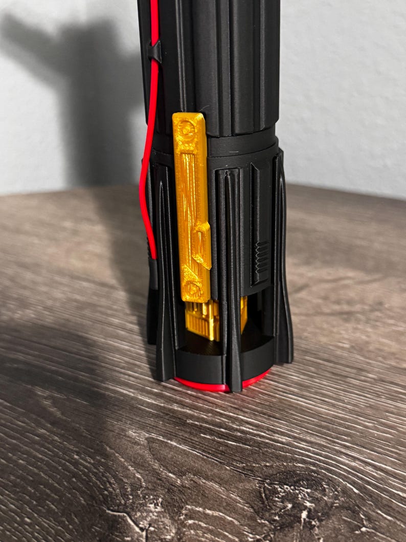 May include: A black and gold lightsaber hilt with red detailing. The hilt has a gold rectangular feature and a red wire. The base is encircled by a red ring.