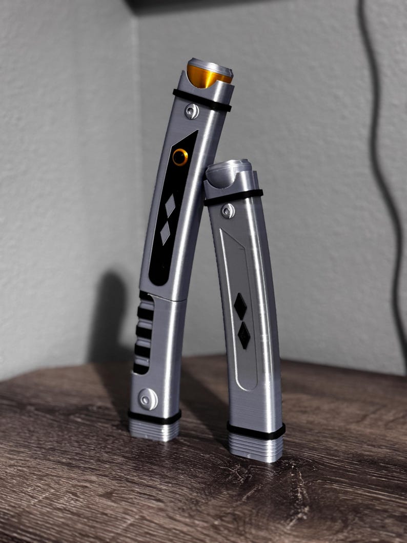 Pu&ograve; includere: Due spade laser argentate con dettagli neri e dorati. La spada laser pi&ugrave; grande presenta un pannello nero con diamanti e scanalature. La spada laser pi&ugrave; piccola ha un design simile. Entrambe hanno fasce nere e sono esposte su una superficie di legno.