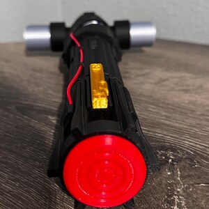 May include: A black and red lightsaber with silver cross-guard blades and a gold accent. The lightsaber has a red circular end and a black handle. The lightsaber is a prop from a science fiction movie.