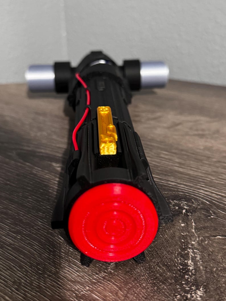 May include: A black and red lightsaber with silver cross-guard blades and a gold accent. The lightsaber has a red circular end and a black handle. The lightsaber is a prop from a science fiction movie.