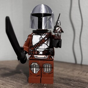 May include: A plastic minifigure featuring a silver helmet, brown and white armor, and a black sword. The figure holds a smaller, matching miniature version of itself in one hand. The design includes detailed printed patterns on the torso and legs.
