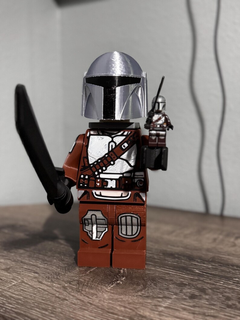 May include: A plastic minifigure featuring a silver helmet, brown and white armor, and a black sword. The figure holds a smaller, matching miniature version of itself in one hand. The design includes detailed printed patterns on the torso and legs.