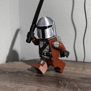 May include: A plastic toy figure resembling a space warrior wearing a metallic silver helmet with a black T-shaped visor. The figure has a brown torso and legs, black hands, and holds a black sword-like weapon raised in its right hand.