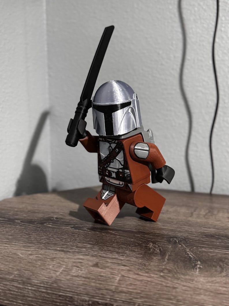 May include: A plastic toy figure resembling a space warrior wearing a metallic silver helmet with a black T-shaped visor. The figure has a brown torso and legs, black hands, and holds a black sword-like weapon raised in its right hand.