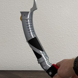 May include: A silver and black lightsaber prop with a curved design. The hilt features a textured grip, a red button, and a yellow accent. The top section has a curved blade-like element. The prop is held by a person.