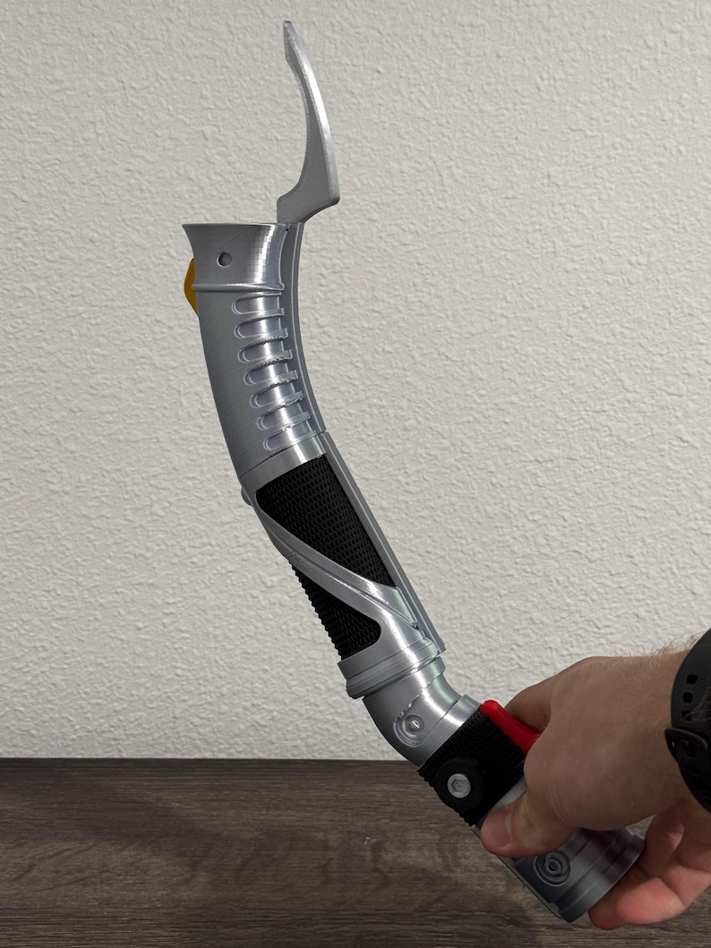 May include: A silver and black lightsaber prop with a curved design. The hilt features a textured grip, a red button, and a yellow accent. The top section has a curved blade-like element. The prop is held by a person.