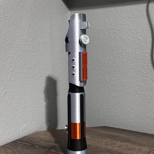 May include: A silver and black lightsaber prop with orange and red accents. The prop has a cylindrical design with detailed features, including buttons and a textured grip. The lightsaber is displayed on a wooden surface.