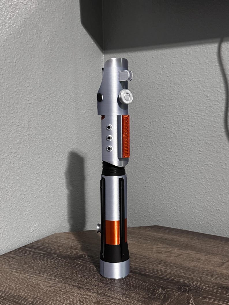 May include: A silver and black lightsaber prop with orange and red accents. The prop has a cylindrical design with detailed features, including buttons and a textured grip. The lightsaber is displayed on a wooden surface.