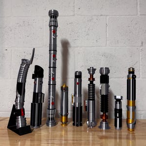 May include: A collection of nine lightsabers with various designs and colors, including silver, black, and gold. Some have red or orange accents. They are arranged on a wooden surface against a white brick wall.