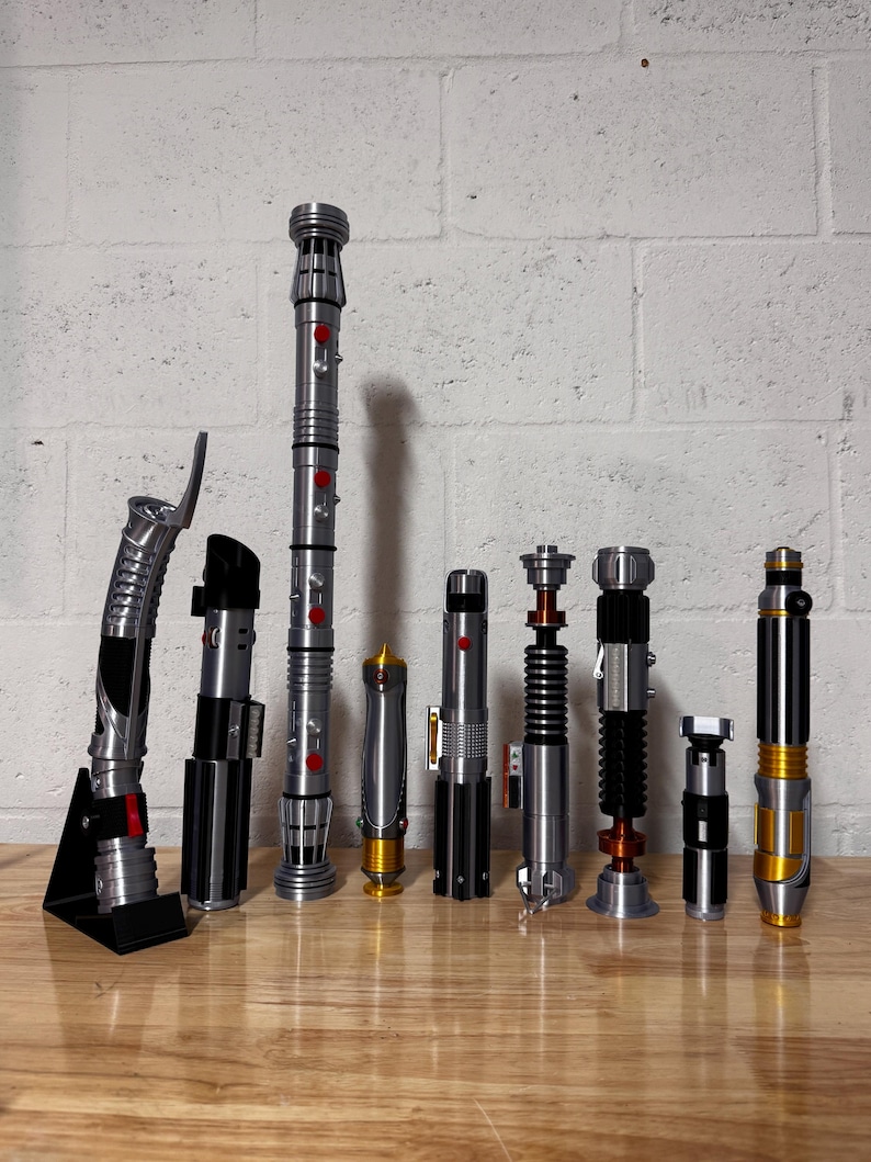 May include: A collection of nine lightsabers with various designs and colors, including silver, black, and gold. Some have red or orange accents. They are arranged on a wooden surface against a white brick wall.