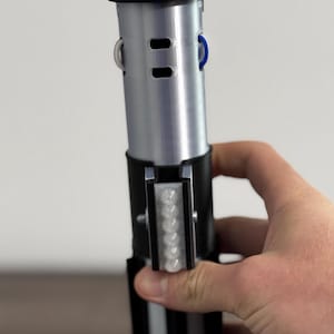 Pu&ograve; includere: Un'elica di spada laser argento e nera con un pulsante blu e una serie di piccole luci. L'elica &egrave; tenuta da una mano e ha un'impugnatura nera con strisce bianche.