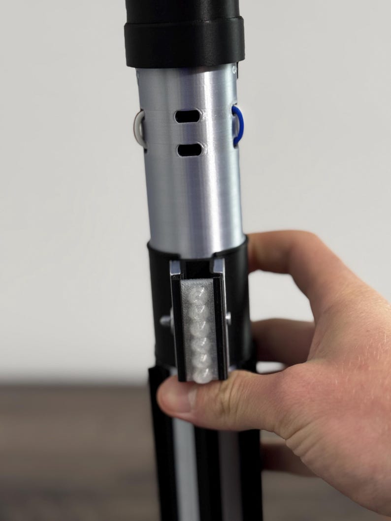 Pu&ograve; includere: Un'elica di spada laser argento e nera con un pulsante blu e una serie di piccole luci. L'elica &egrave; tenuta da una mano e ha un'impugnatura nera con strisce bianche.