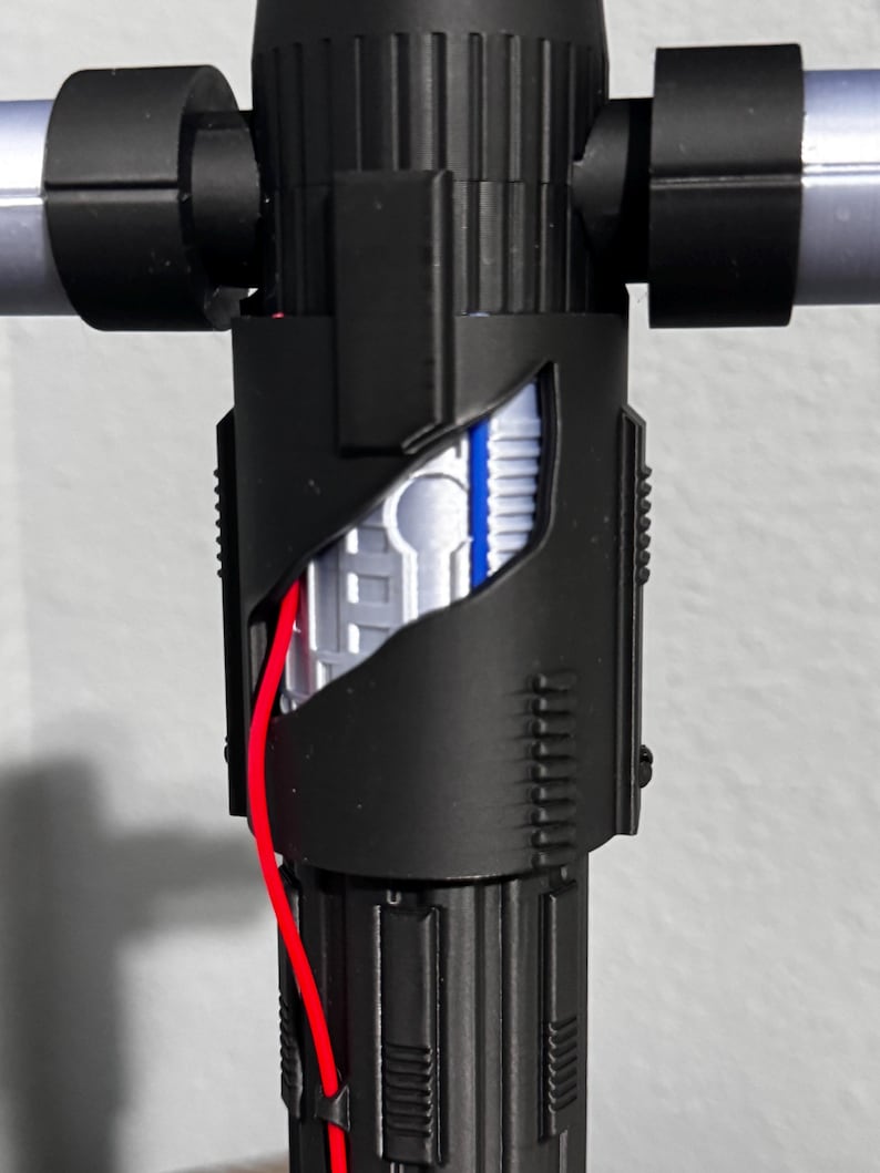 May include: A black lightsaber hilt with silver crossguard arms. The hilt features intricate detailing and a cutaway section showing internal components, including a red wire. This is a prop or toy lightsaber, suitable for cosplay.