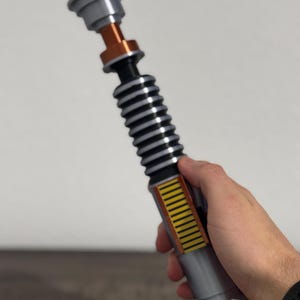 Puede incluir: Un sable de luz plateado y negro con detalles en naranja y amarillo. El accesorio tiene una secci&oacute;n negra acanalada, un mango plateado y una parte superior detallada. El sable de luz se sostiene en una mano.